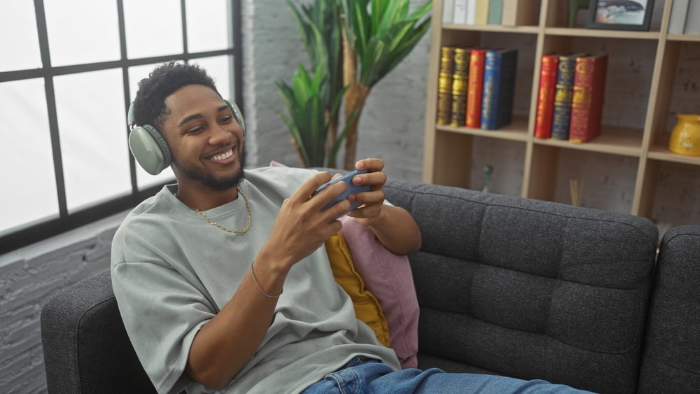 Smiling man playing mobile game on couch indoors with comfortable headphones
