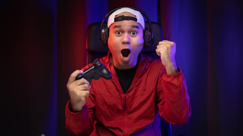 Excited and shocked face of Asian gamer with headphone holding joystick playing video game online sitting on chair at living room. Indian professional gamer streaming on social playing game very fun